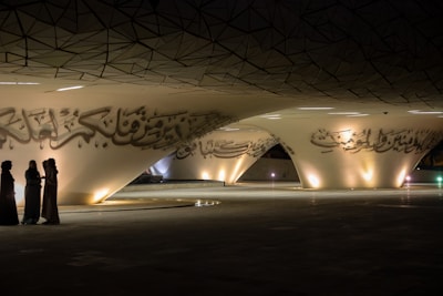 A modern architectural space featuring large, sweeping curves with Arabic calligraphy adorning the surfaces. Three people are standing together in discussion, dressed in traditional Middle Eastern attire. The area is dimly lit with soft, warm lights highlighting the calligraphic details.