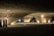 A modern architectural space featuring large, sweeping curves with Arabic calligraphy adorning the surfaces. Three people are standing together in discussion, dressed in traditional Middle Eastern attire. The area is dimly lit with soft, warm lights highlighting the calligraphic details.