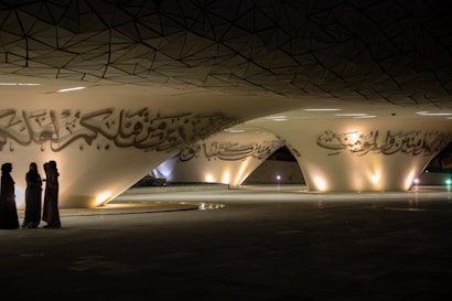 A modern architectural space featuring large, sweeping curves with Arabic calligraphy adorning the surfaces. Three people are standing together in discussion, dressed in traditional Middle Eastern attire. The area is dimly lit with soft, warm lights highlighting the calligraphic details.