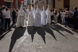 A group of people wearing white robes with tall pointed hoods and carrying staffs are walking down a street. They are surrounded by onlookers. The sun casts long, dark shadows of the hooded figures onto the street. The street is lined with buildings, and some people are taking photos.