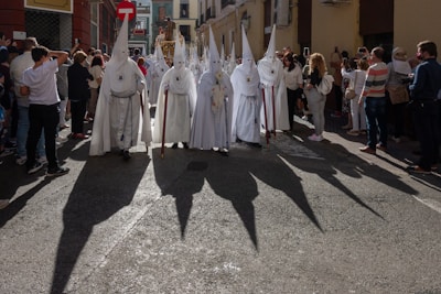 A group of people wearing white robes with tall pointed hoods and carrying staffs are walking down a street. They are surrounded by onlookers. The sun casts long, dark shadows of the hooded figures onto the street. The street is lined with buildings, and some people are taking photos.