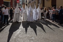 A group of people wearing white robes with tall pointed hoods and carrying staffs are walking down a street. They are surrounded by onlookers. The sun casts long, dark shadows of the hooded figures onto the street. The street is lined with buildings, and some people are taking photos.