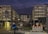 A construction or demolition site surrounded by a chain-link fence, with two large buildings flanking a central area filled with debris and construction machinery. A cyclist wearing a blue jacket and helmet rides past the site in the foreground. The sky is dark, suggesting early morning or evening, and bright lights illuminate the area.