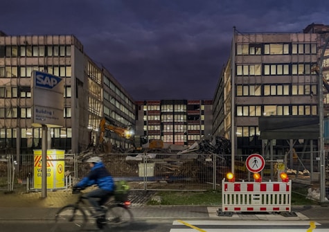 A construction or demolition site surrounded by a chain-link fence, with two large buildings flanking a central area filled with debris and construction machinery. A cyclist wearing a blue jacket and helmet rides past the site in the foreground. The sky is dark, suggesting early morning or evening, and bright lights illuminate the area.
