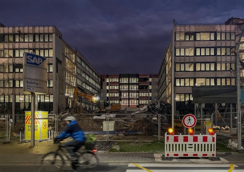 A construction or demolition site surrounded by a chain-link fence, with two large buildings flanking a central area filled with debris and construction machinery. A cyclist wearing a blue jacket and helmet rides past the site in the foreground. The sky is dark, suggesting early morning or evening, and bright lights illuminate the area.