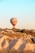 A hot air balloon floats above a rugged and scenic landscape, with sandy-colored hills and sparse vegetation. The balloon has a label that reads 'Flower Balloons' and it stands prominently against a clear blue sky. Sunlight casts warm tones across the terrain, creating a peaceful and serene scene.