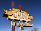 A vintage, weathered sign for a cafe that offers Mexican food. The sign has a distinctly retro look with large, bold letters and an old-fashioned style. The words 'RANCH HOUSE' and 'MEXICAN FOOD' are prominently displayed. The sign is partially rusted and faded, suggesting it has been exposed to the elements for a long time. The background is a clear blue sky.