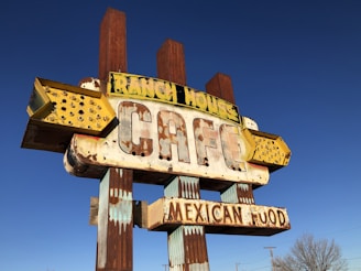 A vintage, weathered sign for a cafe that offers Mexican food. The sign has a distinctly retro look with large, bold letters and an old-fashioned style. The words 'RANCH HOUSE' and 'MEXICAN FOOD' are prominently displayed. The sign is partially rusted and faded, suggesting it has been exposed to the elements for a long time. The background is a clear blue sky.