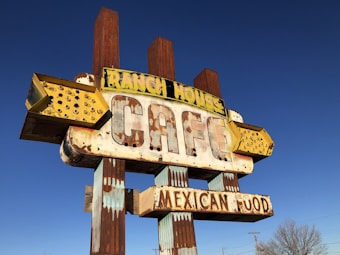 A vintage, weathered sign for a cafe that offers Mexican food. The sign has a distinctly retro look with large, bold letters and an old-fashioned style. The words 'RANCH HOUSE' and 'MEXICAN FOOD' are prominently displayed. The sign is partially rusted and faded, suggesting it has been exposed to the elements for a long time. The background is a clear blue sky.