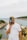 A man stands by a scenic coastal landscape, holding an old-fashioned camera up to his face. He wears a white shirt with the words 'Deus Customs Parts & Service' and a khaki cap. In the background, there are rocky cliffs, lush greenery, and a calm sea under a cloudy sky.