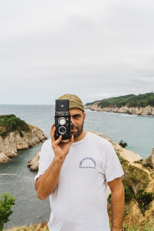 A man stands by a scenic coastal landscape, holding an old-fashioned camera up to his face. He wears a white shirt with the words 'Deus Customs Parts & Service' and a khaki cap. In the background, there are rocky cliffs, lush greenery, and a calm sea under a cloudy sky.