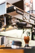 A close-up view of a professional espresso machine in a coffee shop, showcasing various controls and portafilters. Other items such as a tamper and coffee supplies are present on the counter near the machine.