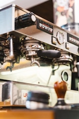 A close-up view of a professional espresso machine in a coffee shop, showcasing various controls and portafilters. Other items such as a tamper and coffee supplies are present on the counter near the machine.