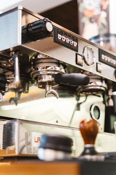 A close-up view of a professional espresso machine in a coffee shop, showcasing various controls and portafilters. Other items such as a tamper and coffee supplies are present on the counter near the machine.