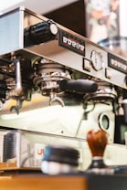 A close-up view of a professional espresso machine in a coffee shop, showcasing various controls and portafilters. Other items such as a tamper and coffee supplies are present on the counter near the machine.