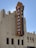 A large, vertical red and yellow sign displaying the word 'Paramount' is prominently attached to the side of an ornate, light-colored building with multiple windows. The structure features an Art Deco architectural style, with decorative elements and embellishments. The sky is clear and blue in the background.