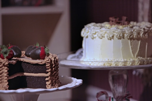 Two cakes on display; one is a chocolate cake with layers covered in chocolate frosting and topped with chocolate-covered strawberries, while the other is a white frosted cake with decorative cream swirls.