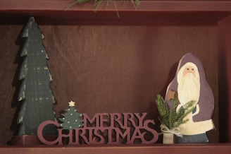A wooden decoration featuring a tall, green Christmas tree and a Santa Claus figure. In front, the words 'Merry Christmas' are cut out in bold, red wooden letters. A small sprig of evergreen is attached to the Santa figure, adding a touch of festivity.
