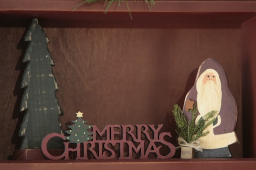 A wooden decoration featuring a tall, green Christmas tree and a Santa Claus figure. In front, the words 'Merry Christmas' are cut out in bold, red wooden letters. A small sprig of evergreen is attached to the Santa figure, adding a touch of festivity.