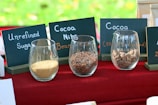 Glass containers placed on a red tablecloth display unrefined sugar, cocoa nibs, and cocoa beans. Behind each container are chalkboard labels describing the contents. The background has a blurred appearance, suggesting an outdoor setting.