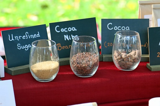 Glass containers placed on a red tablecloth display unrefined sugar, cocoa nibs, and cocoa beans. Behind each container are chalkboard labels describing the contents. The background has a blurred appearance, suggesting an outdoor setting.