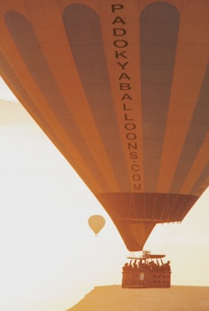 A large hot air balloon with the text 'PADOKYABALLOONS.COM' is seen from beneath, capturing the balloon's expansive stripes and basket carrying several people. Another smaller balloon floats in the background against a backdrop of a soft, golden sky.