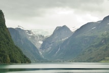 A picturesque landscape featuring a serene lake surrounded by majestic mountains with snow-capped peaks. The lush greenery of the hillsides contrasts with the white of the snow and glacier. The sky is overcast, creating a muted and tranquil atmosphere.