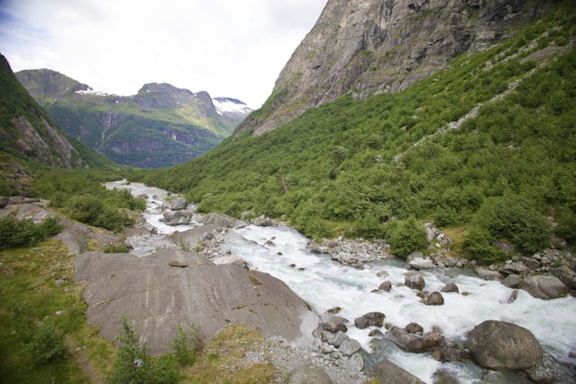 A serene mountain landscape with a river flowing through the valley, surrounded by lush greenery and rocky slopes. The mountains are partially covered with snow in the background, creating a picturesque view.