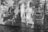 A double exposure black and white photograph featuring historic brick buildings with large windows alongside a canal. Reflections and overlapping images create an artistic and layered effect, with vines and water adding texture.