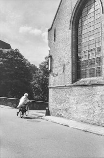 An elderly person is seen riding a bicycle on a cobblestone street next to an old brick building with a large arched window. Trees and another building are visible in the background, under a partly cloudy sky.
