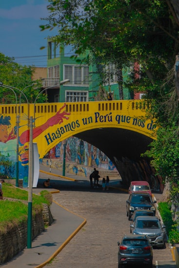 A colorful urban scene featuring a mural on a bridge with vibrant artwork and a message. The bridge is adorned with a yellow banner displaying a motivational phrase in Spanish. Several cars are parked on the street below, and pedestrians are walking. Surrounding the bridge are green trees and multi-story buildings painted in shades of green.