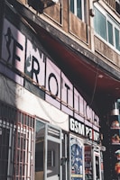 An urban street scene featuring several storefronts. Prominent signage includes a partially visible large faded sign and another that reads 'GSM POINT'. The buildings are aged, with visible wear and discoloration on the walls. There are multiple advertisements and posters in the shop windows and on the doors. A shadow is cast over part of the scene, adding a stark contrast effect.