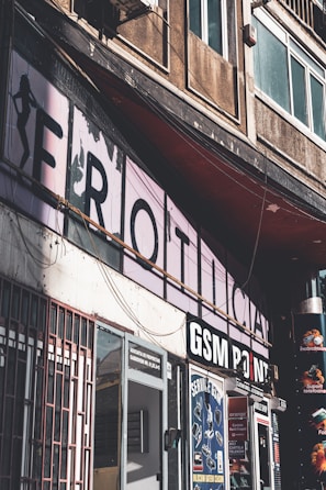 An urban street scene featuring several storefronts. Prominent signage includes a partially visible large faded sign and another that reads 'GSM POINT'. The buildings are aged, with visible wear and discoloration on the walls. There are multiple advertisements and posters in the shop windows and on the doors. A shadow is cast over part of the scene, adding a stark contrast effect.
