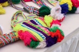 Colorful woven bags with intricate geometric patterns and vibrant tassels are displayed on a table. The bags feature a blend of bright colors, including neon green, red, purple, blue, and white. The design includes symmetrical shapes and stripes, adding to the detailed craftsmanship of the textile work.