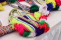Colorful woven bags with intricate geometric patterns and vibrant tassels are displayed on a table. The bags feature a blend of bright colors, including neon green, red, purple, blue, and white. The design includes symmetrical shapes and stripes, adding to the detailed craftsmanship of the textile work.