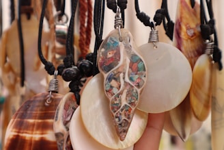 A collection of various shell necklaces hanging closely together, featuring a mix of smooth and polished surfaces. Some shells are circular while one is uniquely leaf-shaped and decorated with colorful embedded stones. The necklaces are suspended on black cords, some with metallic accents.