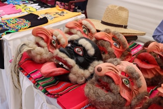 A collection of colorful traditional masks with detailed facial features, such as exaggerated noses and beards, laid out on a vibrant textile. A straw hat is placed nearby, and several brightly colored t-shirts are visible in the background.