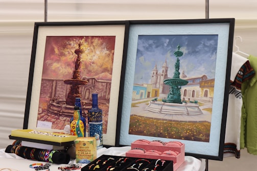 Two framed paintings of ornate fountains are displayed side by side. The left painting features a dark-toned fountain with a fiery, dramatic sky. The right painting depicts a fountain against a backdrop of a light blue sky and historic buildings. Various colorful handcrafted items, including bottles, bracelets, and jewelry, are arranged in front of the paintings, suggesting a marketplace or art fair setting.