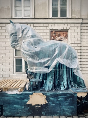 A large sculpture wrapped in protective plastic sits against a building with a brick facade and windows. The sculpture is elevated on a blue-green painted platform, next to which is a wooden pallet and a disposable cup. An inset relief of a reclining figure is integrated into the wall behind.