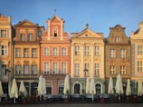 A row of five adjacent, colorful historical buildings, each with unique architectural details and designs. The facades have intricate moldings, decorative elements, and attic windows under the roofline. In front of the buildings are closed outdoor cafe umbrellas, suggesting a quiet, early morning or evening setting.