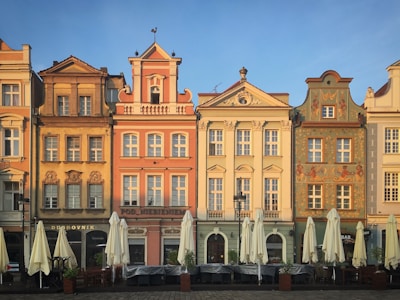 A row of five adjacent, colorful historical buildings, each with unique architectural details and designs. The facades have intricate moldings, decorative elements, and attic windows under the roofline. In front of the buildings are closed outdoor cafe umbrellas, suggesting a quiet, early morning or evening setting.