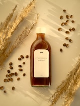 A bottle of cold brew coffee is placed on a surface surrounded by scattered coffee beans and dry wheat stalks. The label on the bottle reads 'COLD BREW BLEND COFFEE' and contains 100 ML. The scene is set in warm lighting, creating a cozy and inviting ambiance.