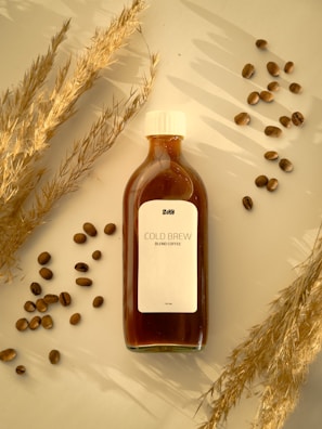 A bottle of cold brew coffee is placed on a surface surrounded by scattered coffee beans and dry wheat stalks. The label on the bottle reads 'COLD BREW BLEND COFFEE' and contains 100 ML. The scene is set in warm lighting, creating a cozy and inviting ambiance.