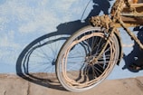 A bicycle with its frame and wheels wrapped in ropes and seashells, casting a shadow on a textured blue wall. It appears weathered and artistic, with intricate details on the ropework and natural elements like barnacles attached. The tires and metal parts are still visible underneath the decorative material.