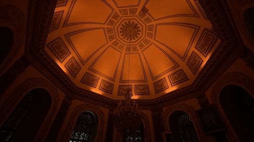 An ornately designed ceiling with intricate geometrical patterns and detailed motifs. The ceiling features warm orange lighting that highlights its architectural elements. A large chandelier hangs prominently in the center, adding an elegant touch to the ambiance. The surrounding walls are adorned with decorative arches and windows.