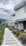 A modern house with a stone facade and a neatly landscaped garden. The garden features a pathway lined with small bushes and various plants, leading up to the entrance. The background includes another residential building and a hill under a cloudy sky.