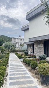 A modern house with a stone facade and a neatly landscaped garden. The garden features a pathway lined with small bushes and various plants, leading up to the entrance. The background includes another residential building and a hill under a cloudy sky.