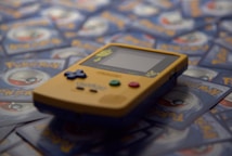 A yellow Game Boy Color handheld gaming console is positioned on top of a pile of Pok&eacute;mon trading cards. The Game Boy features colorful buttons and a small screen. The Pok&eacute;mon cards are blue and gray with a circular emblem in the center.