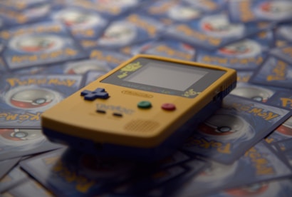 A yellow Game Boy Color handheld gaming console is positioned on top of a pile of Pokémon trading cards. The Game Boy features colorful buttons and a small screen. The Pokémon cards are blue and gray with a circular emblem in the center.