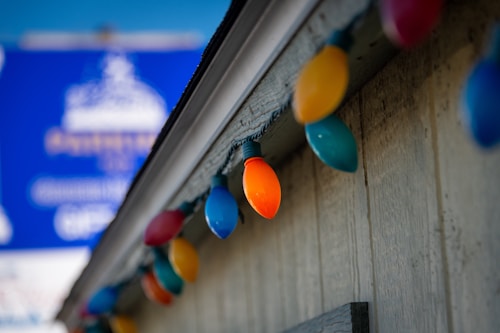 A row of colorful Christmas lights is attached to the edge of a building with a weathered gray wood exterior. The lights are in various colors, including red, yellow, and blue. In the background, there is a blue sign with indistinct white and yellow text.